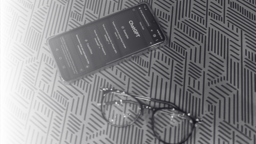 Black framed reading glasses sit on a desk next to a mobile phone which is switched on and showing the website of the AI tool Chap GPT
