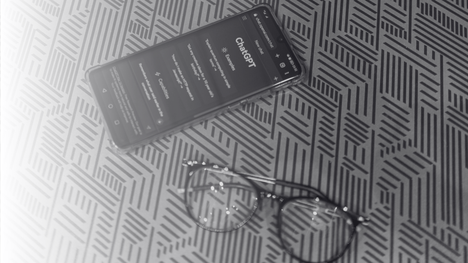 Black framed reading glasses sit on a desk next to a mobile phone which is switched on and showing the website of the AI tool Chap GPT